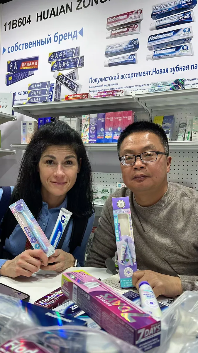 A Russian buyer and the Huai'an Zongheng export representative holding private label toothpaste samples at the factory's trade show stand, with the booth number 11B604 and Russian product signage visible behind them